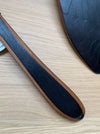 A close-up of a dark wood-handled kitchen utensil, likely a spoon or spatula, resting on a light wood grain surface. The handle features a dark black center with a warm brown wood grain border, and another matching dark utensil is partially visible in the upper right.