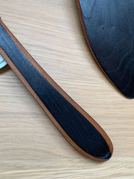 A close-up of a dark wood-handled kitchen utensil, likely a spoon or spatula, resting on a light wood grain surface. The handle features a dark black center with a warm brown wood grain border, and another matching dark utensil is partially visible in the upper right.