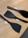 Three black wooden spatulas with light brown wood edges, arranged diagonally on a light wood grain tabletop. Each spatula has a small white paper tag attached near the handle.