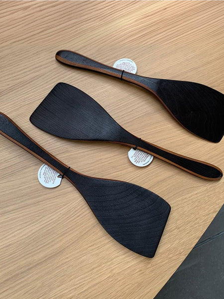 Three black wooden spatulas with light brown wood edges, arranged diagonally on a light wood grain tabletop. Each spatula has a small white paper tag attached near the handle.
