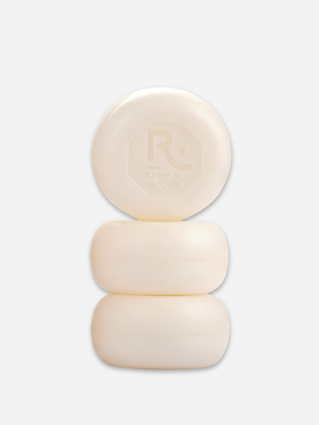 Three round cream-colored bath bars stacked vertically on a light gray background, with the top bar embossed featuring a logo