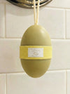 A lavender and rose body butter soap bar in the shape of an egg, hanging from a metal hook on a white tiled wall. The soap is olive green with a yellow horizontal band wrapped around the middle, featuring a white label with gold text.