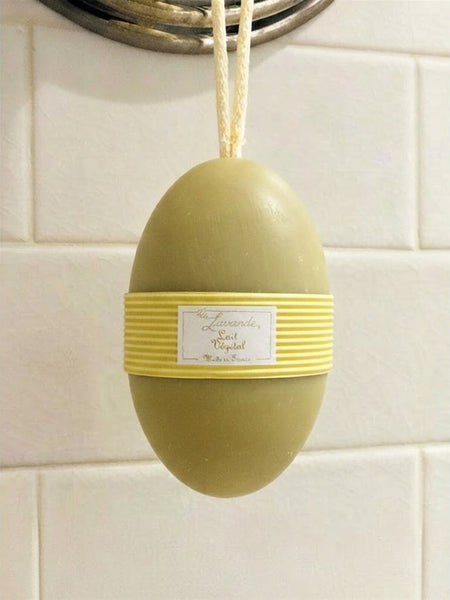 A lavender and rose body butter soap bar in the shape of an egg, hanging from a metal hook on a white tiled wall. The soap is olive green with a yellow horizontal band wrapped around the middle, featuring a white label with gold text.