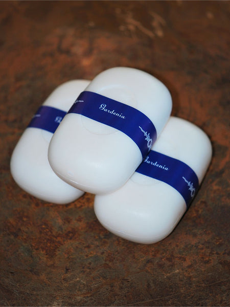 Three white oval-shaped soap bars wrapped in blue paper bands with the brand name Gardennie, arranged on a rustic brown textured surface.