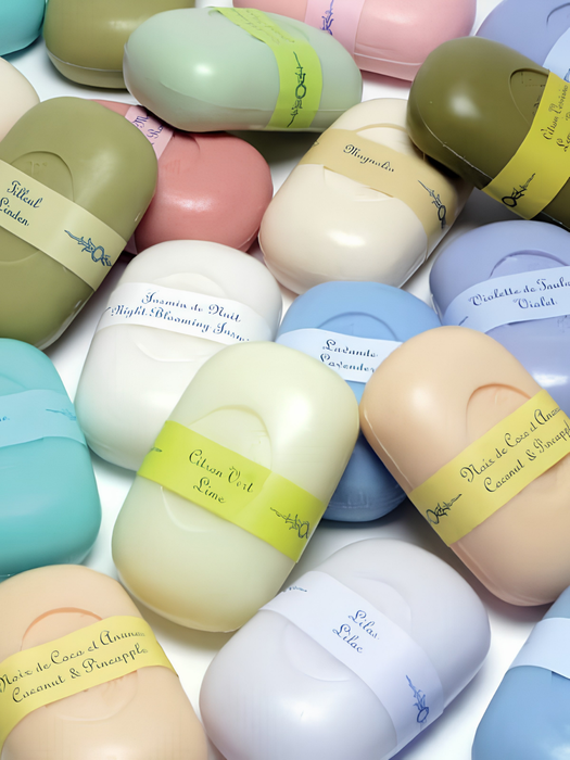 A close-up arrangement of small oval cosmetic or bath product containers in pastel colors, each wrapped with a yellow band and labeled with scents such as Sandalwood, Jasmine Night Melody, Lavender, and Coconut & Pineapple.