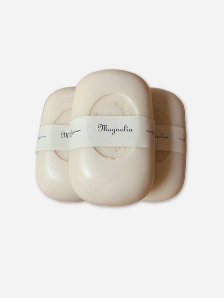 Three oval-shaped cream-colored soap bars wrapped in white paper bands labeled 'Magnolia', arranged in a fan-like composition on a light gray background.