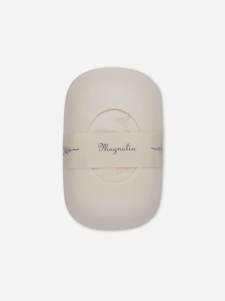 An oval-shaped cream-colored soap bar wrapped in a beige paper band labeled 'Magnolia' on a light gray background.