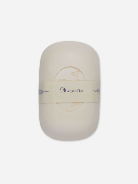 An oval-shaped cream-colored soap bar wrapped in a beige paper band labeled 'Magnolia' on a light gray background.