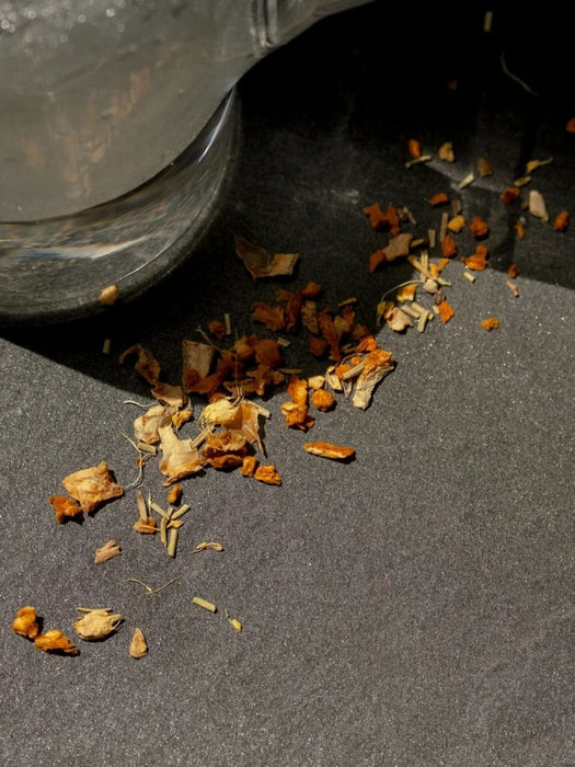 Dried orange and beige flower petals scattered on a dark gray surface beneath the edge of a clear glass container.