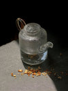 A small clear glass teapot with a loop handle and spout, shown on a dark surface with dried tea leaves scattered in front, against a black background.