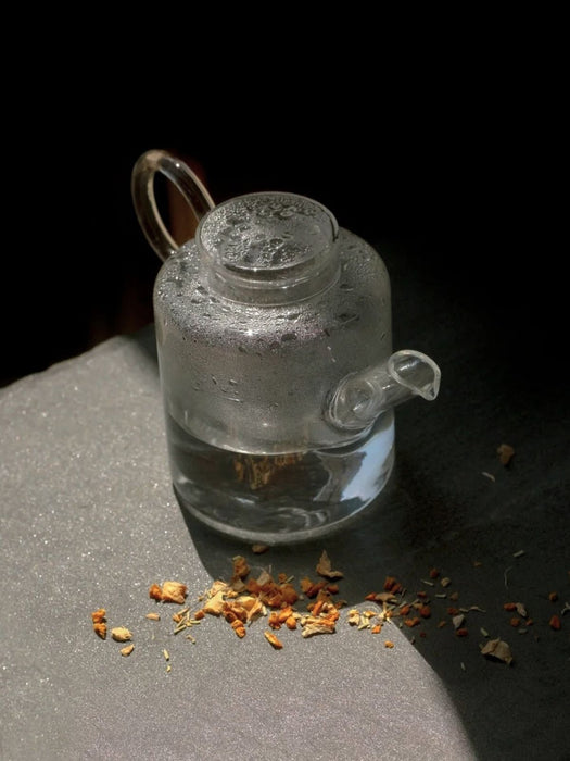 A small clear glass teapot with a loop handle and spout, shown on a dark surface with dried tea leaves scattered in front, against a black background.