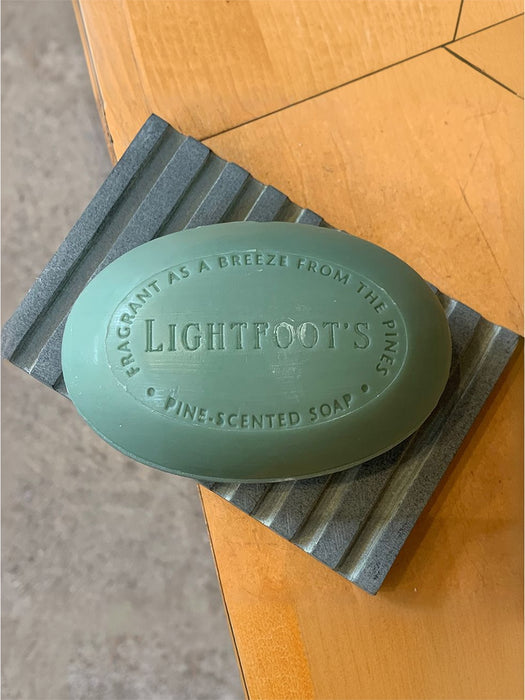 A light green oval bar of Lightfoot's Pines Scented Soap resting on a gray textured soap dish over a wooden table.