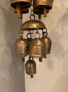 A rustic brass wind chime featuring a large dome-shaped top bell with five smaller hanging bells below, suspended against a textured off-white wall.