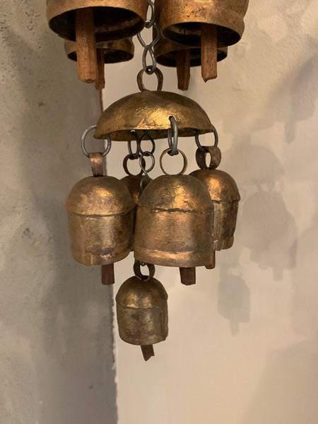 A rustic brass wind chime featuring a large dome-shaped top bell with five smaller hanging bells below, suspended against a textured off-white wall.