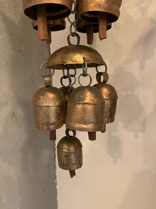 A rustic brass wind chime featuring a large dome-shaped top bell with five smaller hanging bells below, suspended against a textured off-white wall.