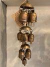 A rustic three-tier wind chime featuring small bronze-colored metal bells with a weathered, antique finish, hanging against a textured off-white wall.
