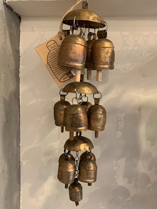 A rustic three-tier wind chime featuring small bronze-colored metal bells with a weathered, antique finish, hanging against a textured off-white wall.