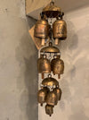 A hanging wind chime made of multiple bronze-toned metal bells of varying sizes, suspended against a textured off-white wall.