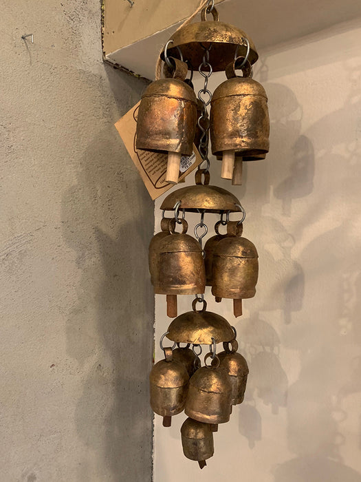 A hanging wind chime made of multiple bronze-toned metal bells of varying sizes, suspended against a textured off-white wall.