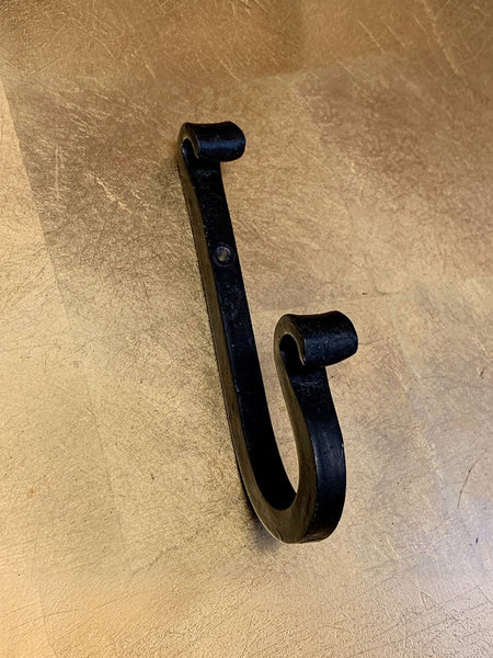 A black cast iron J-hook wall hook with a rustic, hand-forged texture, shown on a light brown wood-grain surface.