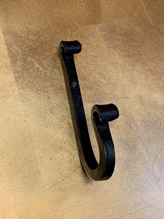 A black cast iron J-hook wall hook with a rustic, hand-forged texture, shown on a light brown wood-grain surface.