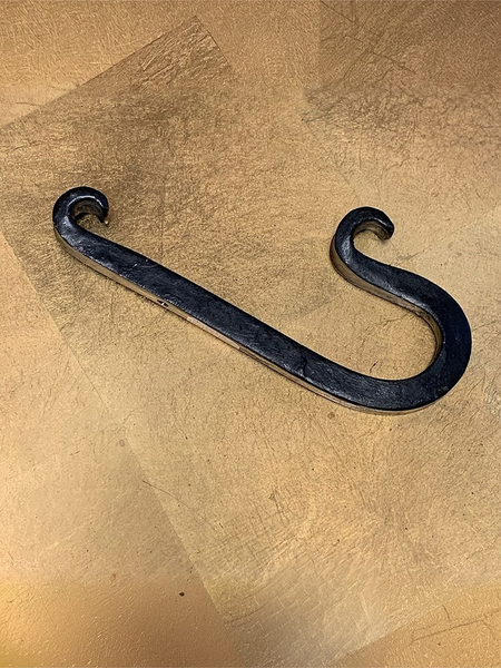 A dark metal J-shaped hook with a textured, hand-forged finish, resting on a textured gold surface.