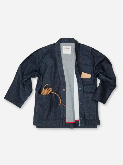 A dark indigo denim jacket from the brand Kith, styled open over a gray knit sweater. The jacket features a collar, front button closure, a chest pocket with a beige leather-like accent, and a large front pocket with a coiled tan rope detail, all laid flat on a white background.