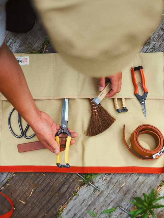 A top-down view of a person's hands organizing gardening tools on a beige canvas roll-up tool bag with an orange-red border. The tools include yellow-handled shears, orange-handled pruning shears, a brown brush, and a brown leather whip, laid out on a weathered wooden deck.