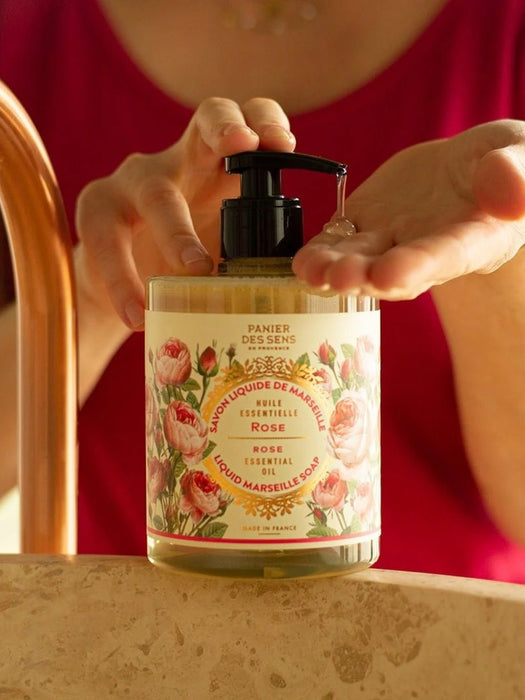 A person at a bathroom counter pumps liquid hand soap from a clear bottle with a black pump. The label reads 'Panier des Sens' and 'Rose'.