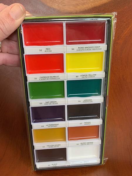A hand holds a 12-color watercolor paint palette in its packaging, showing various paint colors like red, yellow, green, and purple, set against a wooden background.