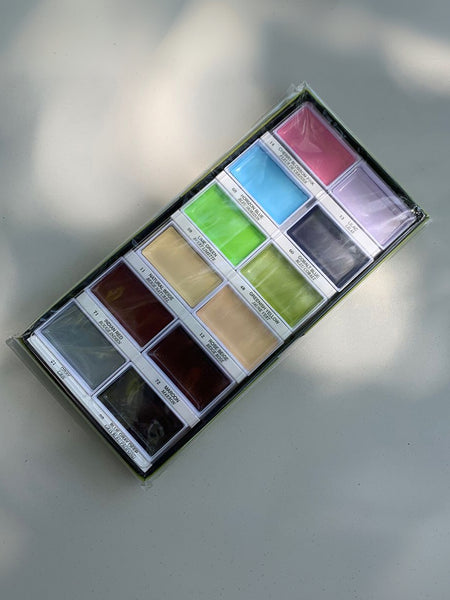 A 16-color acrylic paint palette in a clear plastic case, photographed from an overhead angle on a light gray surface with soft shadows.
