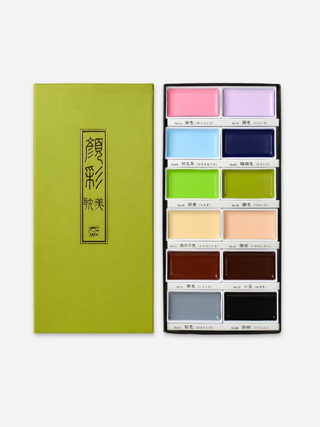 A green rectangular box with gold Chinese characters stands beside an open palette containing multiple color pans, including pink, purple, blue, navy, green, lime, beige, peach, brown, gray, and black, on a light gray background.