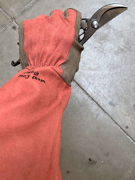 A close-up of a person's hand wearing a long orange-red leather gardening glove with brown trim and black 'West Coast Blades' branding, holding pruning shears over a gray concrete floor.