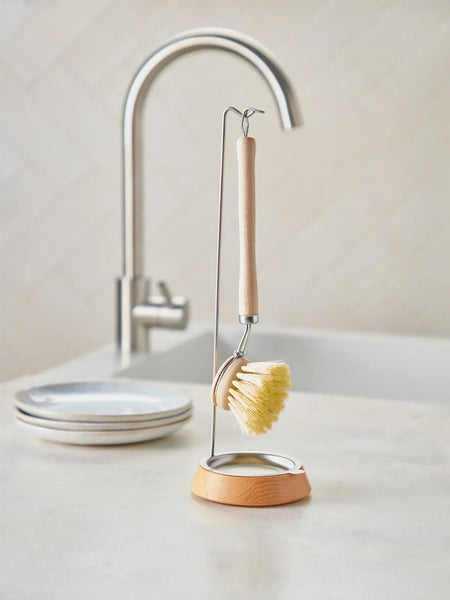 A wooden-handled dish brush with a metal loop and stand, displayed on a light countertop in front of a sink and faucet. A stack of white plates is blurred in the background.