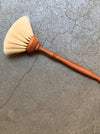 A hand-painted wooden brush with a warm reddish-brown handle and cream-colored bristles, resting diagonally on a textured gray concrete surface.