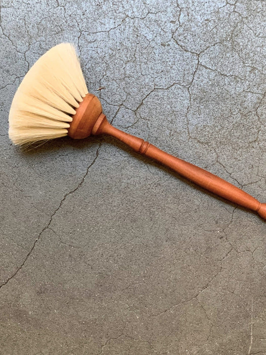 A hand-painted wooden brush with a warm reddish-brown handle and cream-colored bristles, resting diagonally on a textured gray concrete surface.