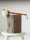 A handmade dust brush with a white bristle head and a curved brown wooden handle, resting on top of a rustic wooden block.
