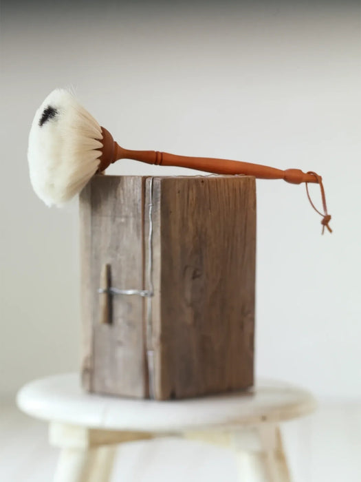 A handmade dust brush with a white bristle head and a curved brown wooden handle, resting on top of a rustic wooden block.