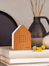 A house-shaped wooden speaker with a white side panel sits on stacked books in a styled interior setting, with a dark vase and dried stems in the background.