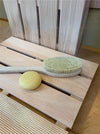 A wooden bath brush with a pale yellow bar of soap resting on its slatted wooden stand, set against a background of matching wooden panels.