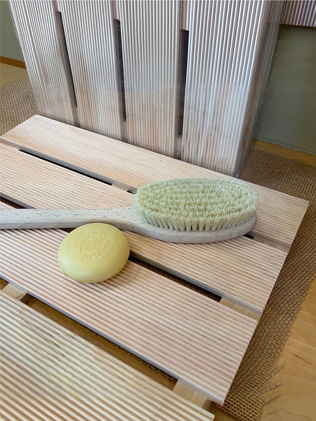 A wooden bath brush with a pale yellow bar of soap resting on its slatted wooden stand, set against a background of matching wooden panels.
