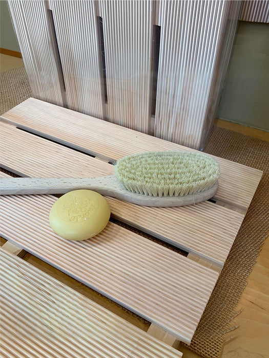A wooden bath brush with a pale yellow bar of soap resting on its slatted wooden stand, set against a background of matching wooden panels.
