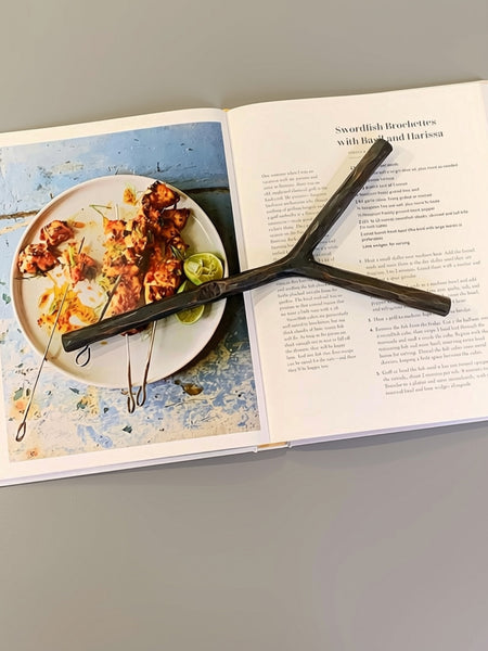 An Iron Y-shaped Metal Bookmark resting on an open cookbook, with a plate of grilled skewers pictured on the left page.