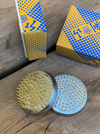 Two round metal nail-hammering tools with gold and silver spikes, displayed on a rustic wooden surface with their blue and yellow patterned packaging boxes in the background.