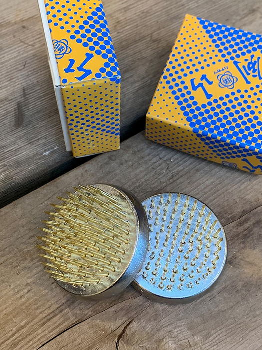 Two round metal nail-hammering tools with gold and silver spikes, displayed on a rustic wooden surface with their blue and yellow patterned packaging boxes in the background.