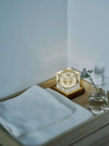 A small illuminated square glass candle holder lit from below with a dandelion inside, sitting on a wooden tray next to folded white towels and a glass of water in a minimalist room corner.