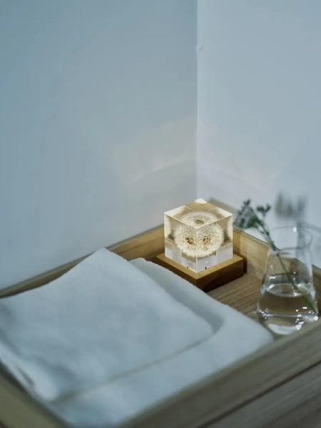 A small illuminated square glass candle holder lit from below with a dandelion inside, sitting on a wooden tray next to folded white towels and a glass of water in a minimalist room corner.