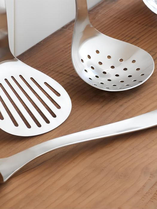 A stainless steel kitchen utensil set including a slotted spoon, a slotted spatula, and a long-handled tool, arranged on a wooden tabletop.