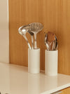 Two white cylindrical utensil holders on a white countertop against a light wood cabinet door, holding stainless steel and wooden kitchen utensils.