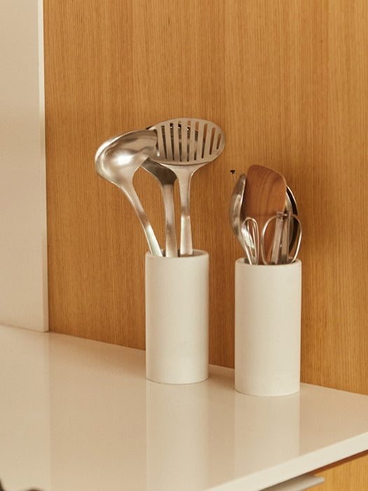 Two white cylindrical utensil holders on a white countertop against a light wood cabinet door, holding stainless steel and wooden kitchen utensils.